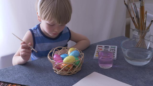 Little blond kid boy coloring eggs for Easter holiday in bath room, indoors. Child holding basket with painted eggs. Child having fun and celebrating feast with easter toy bynny