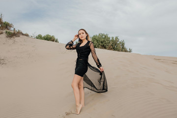 Mujer elegante parada sobre la arena con vestido negro en la playa - obrazy, fototapety, plakaty