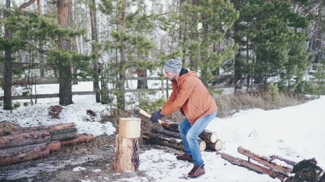 A Caucasian man in a brown jacket, hat, jeans and boots, holding an axe, chopping wood in winter. A man stands in the courtyard of a country house, which is located next to the forest.