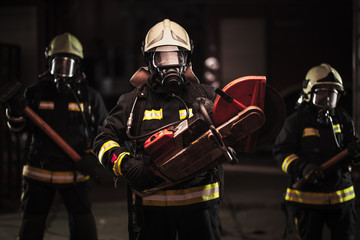 Group of professional firefighters wearing full equipment, oxygen masks, and emergency rescue tools, circular hydraulic and gas saw, axe, and sledge hammer. Firetrucks in the background.