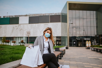 A young beautiful caucasian woman with a mask and bags is sitting on a bench outside the mall. Virus Corona Protection