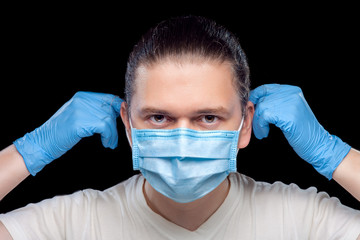 Portrait of a male doctor putting a medical mask on his face with hands in styry gloves, closeup of a medical worker isolated on black background.