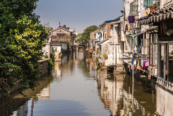 Fototapeta premium Suzhou China - May 3, 2010: City canal with brown water reflects the backside of houses along the water, and green foliage on other side under blue sky. Laundry add colors.