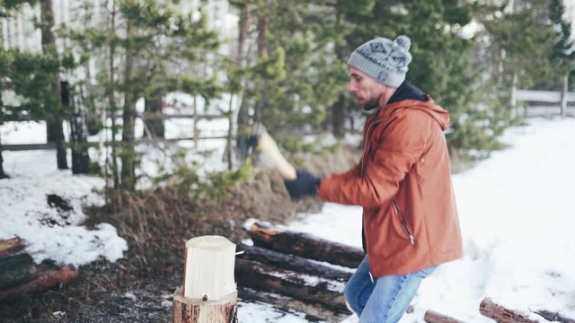 A Caucasian man in a brown jacket, hat, jeans and boots, holding an axe, chopping wood in winter. A man stands in the courtyard of a country house, which is located next to the forest.