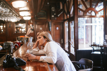 A young attractive caucasian blonde girl with a drink in her hand sits at a bar