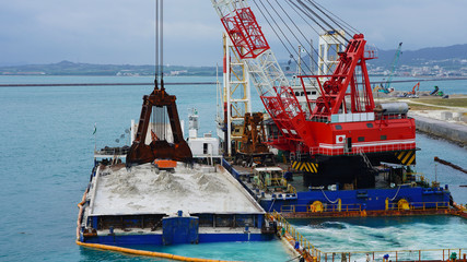 Huge excavator on floating platform in the blue water of the Pacific Ocean extract white sand from the bottom and load onto a barge amid clouds and tropical islands. close up crane basket plunge