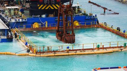 Fototapeta na wymiar Huge excavator on floating platform in the blue water of the Pacific Ocean extract white sand from the bottom and load onto a barge amid clouds and tropical islands. close up crane basket plunge