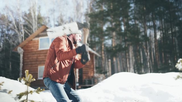A Caucasian man in a brown jacket, hat, jeans and boots, holding an axe, chopping wood in winter. A man stands in the courtyard of a country house, which is located next to the forest.