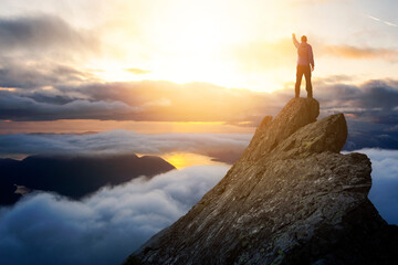 Adventurous Man Hiker With Hands Up on top of a Steep Rocky Cliff. Sunset or Sunrise. Landscape...