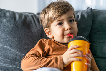 Portrait of small cute caucasian boy three or four years old sitting on the bed or sofa at home holding a plastic cup with straw drinking juice or soda at home alone in day looking to side half length