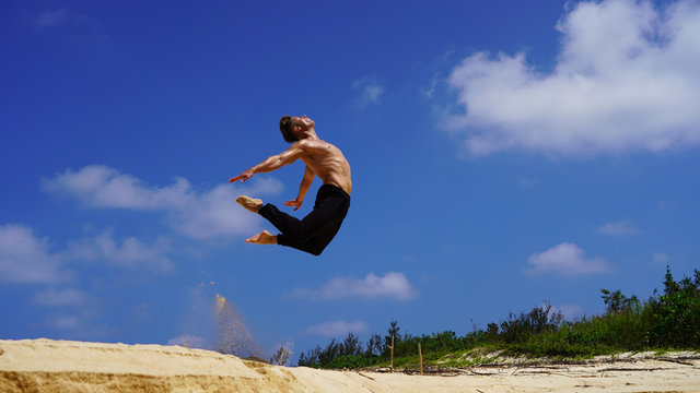 young sporty european guy jumping acrobatics twine. muscular man on the beach shows flexibility back in jumping on background blue sunny sky. stretching