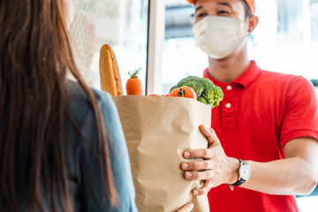 Asian deliver man wearing face mask in red uniform handling bag of food, fruit, vegetable give to female costumer in front of the house. Postman and express grocery delivery service during covid19.