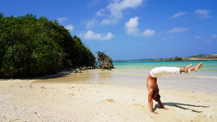 Fototapeta na wymiar muscular acrobat in white pants on the ocean posing standing on his hands in the trough. mexican hand stand on a sunny day on a tropical beach, back band pose