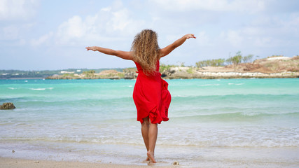 Happy girl in a red bright dress walks along the ocean. Curly woman on vacation on a tropical island