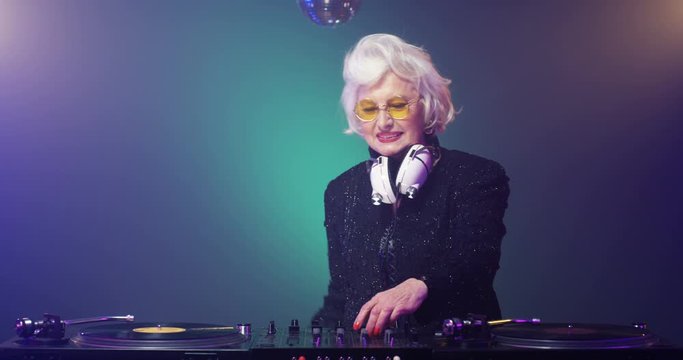 Retro style grey-haired funny Caucasian old female DJ mixing music on audio equipment at club. Stylish vintage senior lady smiling and dancing while playing disco. Clubbing granny. 50s concept