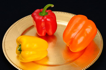 Three bell peppers yellow orange red on a gold plate on black cloth