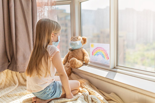 Kid with painted rainbow during Covid-19 quarantine at home. Caucasian Girl with teddy bear toy near window. Stay at home Social media campaign for coronavirus prevention, let's all be well
