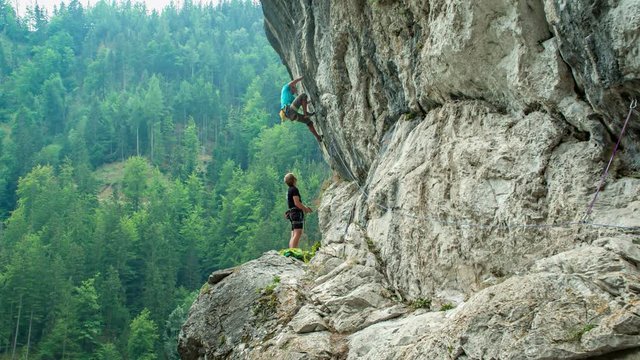 Two men climbing on a high steep wall in the mountains, with spruce forest in the background.