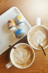 Happy Easter holiday in a cafe, two white cups of coffee on a wooden table, a white plate with colored eggs