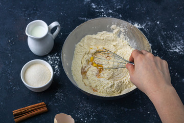 Female hands preparing dough and whisking flour and eggs. Ingredients and utensils for cooking cake (flour, egg, milk, sugar, rolling pin, towel) on dark table. The concept of making dough for baking