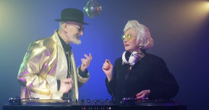 Vintage stylish senior man in sunglasses and lady with headphones dancing near audio equipment. Funny retro style Caucasian couple of old male and female DJs playing disco. 50s party concept