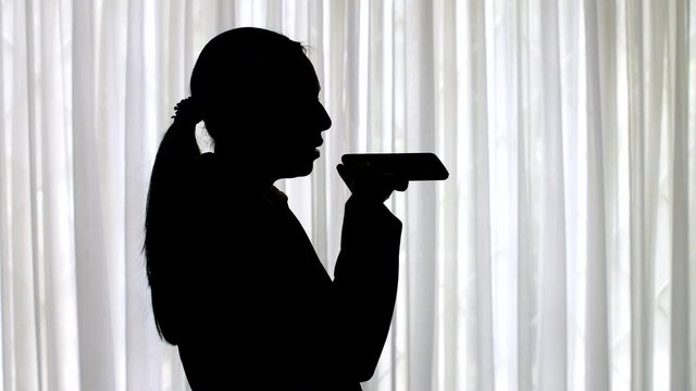 Silhouette of a businesswoman standing in front of a window with curtains, recording an audio voice message on her cellphone