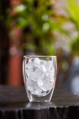 Glass with ice cubes on wooden table with green nature background.