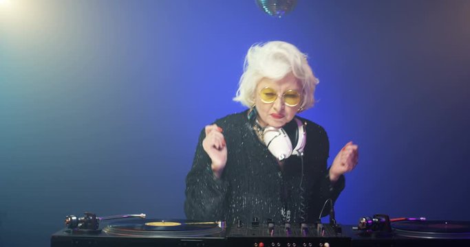 Clubbing grandma DJ dancing while playing disco on dj mixer. Vintage joyful grey-haired lady with headphones at club. Retro style funny Caucasian old pensioner mixing music at party. 60s concept