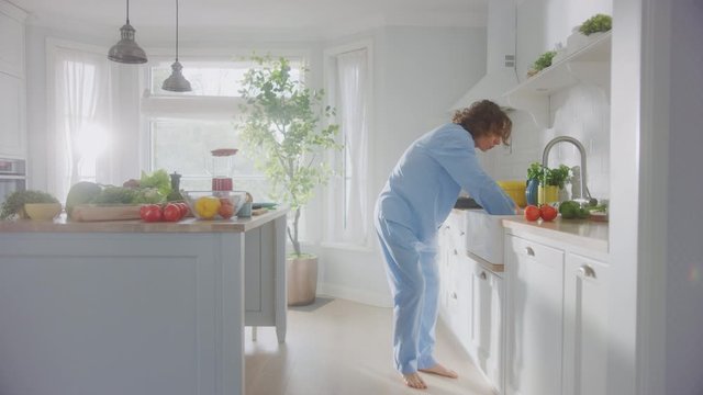 Energetic Funny Young Man with Long Hair Dancing in the Kitchen while Wearing Blue Pajamas. He&rsquo;s Preparing a Salad in a Modern Kitchen Area with Healthy Green Vegetables on a Table. Cozy Home.