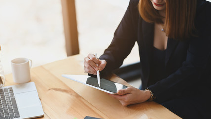 Photo of sexy businesswoman holding a computer tablet and stylus pen while sitting at the wooden working desk over comfortable sitting room as background.