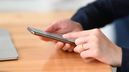 Fototapeta premium Photo of young woman's hands holding and using a smartphone while sitting/relaxing at the wooden working desk over comfortable living room as background.