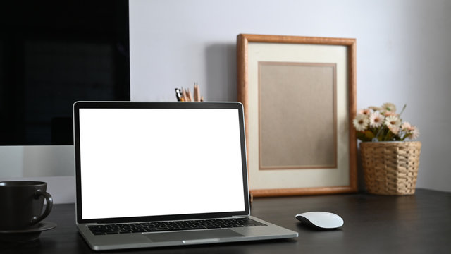 Photo of black working desk along with white blank screen computer laptop, books, notebook, pencil holder, picture frame, potted plant putting together on it with white cement wall as background.
