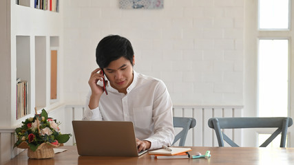 Photo of smart programmer talking on his phone while sitting and using a computer laptop at the wooden working desk over comfortable living room as background.