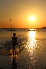 silhouette of person in the foreground and backlight running in the sea at low tide at sunset with boats and promontory in the background
