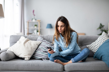 Fototapeta premium Smiling woman relax sit on sofa at home hold cellphone. Online shoping.