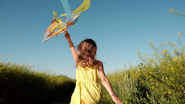 A girl is running across the field in yellow dress with a kite flying over her head. Concept of happy childhood and summer holiday