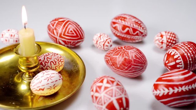 Closeup Red easter eggs with folk Ukrainian pattern lay around and on candlestick with burning candle. And man rotate one egg on white background. Ukrainian traditional eggs pisanka and krashanka