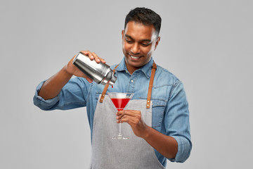alcohol drinks, people and profession concept - indian barman in apron pouring cocktail from shaker to glass over grey background