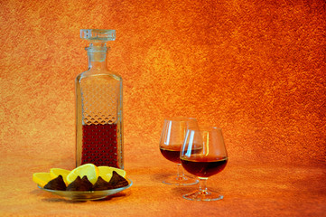 Two glasses and a crystal bottle with cognac and a plate with slices of lemon and chocolate candies...