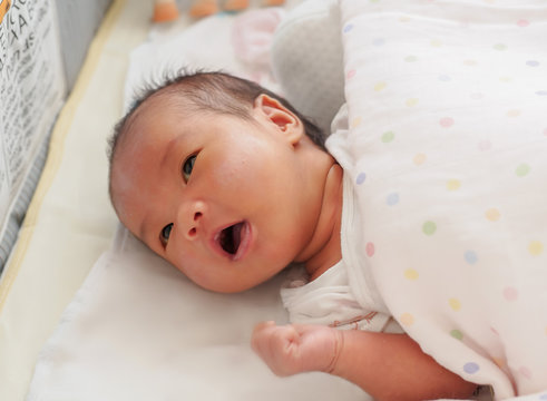 An Asian Newborn Little Girl Is Lying Down On Her Baby Cot And Looking At Her Mother. She Has Some Rash On Her Face, Swaddle With Warm Cloth And Blanket On Top. She Is A Little Open Mouth. It Seems Li