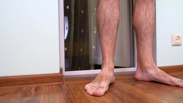 Dowdy man with hairy legs in shorts barefoot crosses the threshold, enters the balcony of a Flat, scratches his leg. Dolly Shot, Moving Camera.