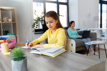 children, education and learning concept - student girl with book writing to notebook and mother working at home