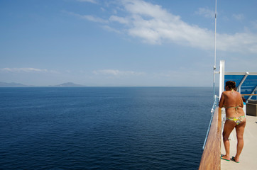 Woman in bikini on deck of luxury cruiseship or cruise ship liner looks towards islands at sea with...