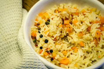 Rice with carrots and onions and black pepper in a white ceramic bowl with textured kitchen towel on background. A protein-free meal of cereals and vegetables.