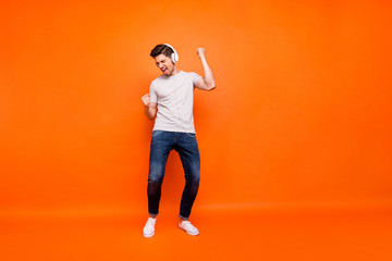 Fototapeta na wymiar Full length photo of funky guy party mood chilling listen modern technology earphones raise hands dancing wear striped t-shirt jeans shoes isolated bright orange color background