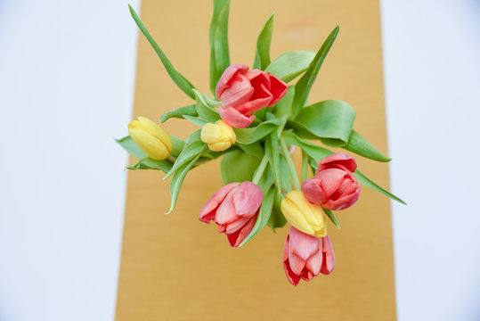 Vertical top down image of a multi colour seven red and yellow Tulips in a vase placed on top of the table with a yellow cover on white surface as a blurred background