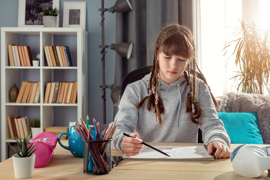 Pensive Female Teenager Studying From Home Sitting At Table Looking At Drawing-book, Backlit Window