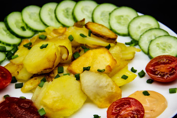 Homemade fried potatoes with fresh cucumbers, cherry tomatoes and sauces on the white plate. Delicious ukrainian fried potatoes with vegetables against background. Cooked tasty meal for lunch.