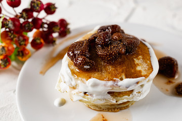 A stack of crumpets and pancakes with sour cream and strawberry sweet jam with berries on top of the dish. Dessert on a white plate with fruit in the background.