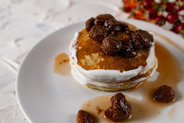 A stack of crumpets and pancakes with sour cream and strawberry sweet jam with berries on top of the dish. Dessert on a white plate with fruit in the background.